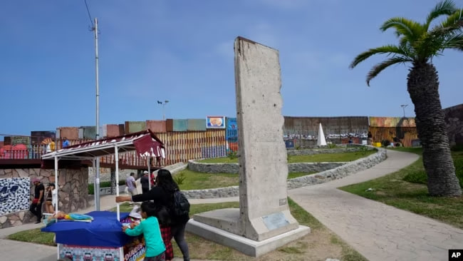 Una familia empuja un carrito de bocadillos más allá de una losa del Muro de Berlín, exhibida cerca del muro fronterizo que separa a Estados Unidos de México, en Tijuana, México, el viernes 25 de agosto de 2023.